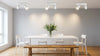 A minimalist dining room with a white table, three white chairs, and a wooden bench. A vase of green foliage centers the table. Soft, warm lighting creates a calm ambiance.