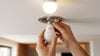 Hands are installing a smart LED bulb into a recessed ceiling socket. The room is softly lit, creating a warm, functional atmosphere.