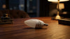 A candle-shaped smart bulb lies on a wooden table in a dimly lit, cozy room, with a blurred view of city lights through the window in the background.